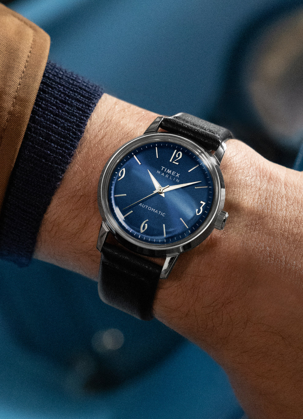 A stylish wrist shot of the watch worn with a brown jacket and navy sweater. The blue sunburst dial catches the light, emphasizing the sleek silver details.