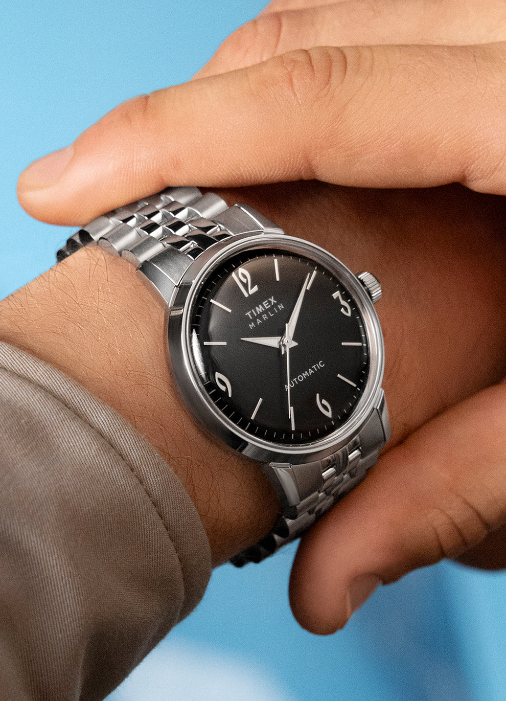 A stylish wrist shot of the black-dial Marlin Automatic, worn with a neutral outfit against a light blue background. The polished case and bracelet contrast with the dark dial.