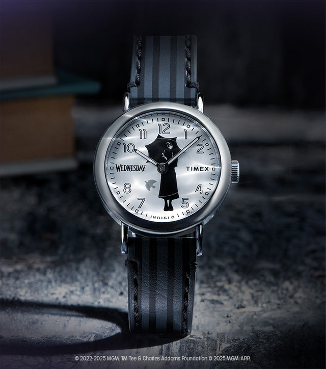 Flatlay of the Timex x Wednesday watch resting on a dark surface with a blurred book in the background, evoking a mysterious, academic aesthetic.