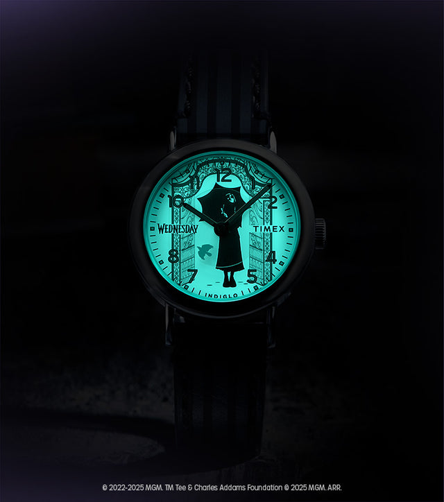 Flatlay of the Timex x Wednesday watch with its Indiglo backlight glowing green in the dark, revealing gothic gate artwork around the dial.