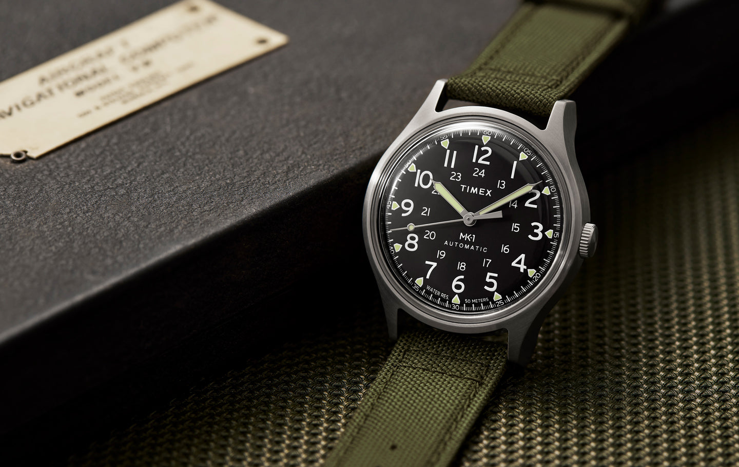 A Timex MK1® Automatic field watch with a black dial, white Arabic numerals, and luminous hands, displayed on an olive green fabric strap. The watch has a stainless steel case and is set against a textured dark background with a metal nameplate. The design reflects a military-inspired aesthetic with clear, easy-to-read markings and a rugged yet stylish appearance.