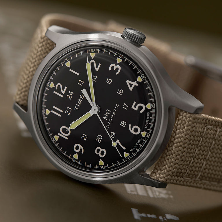 Close-up view of a Timex MK1 Automatic watch with a black dial featuring both 12-hour and 24-hour numerals, luminous hands, and a beige fabric strap. The watch is set against a blurred olive green background.