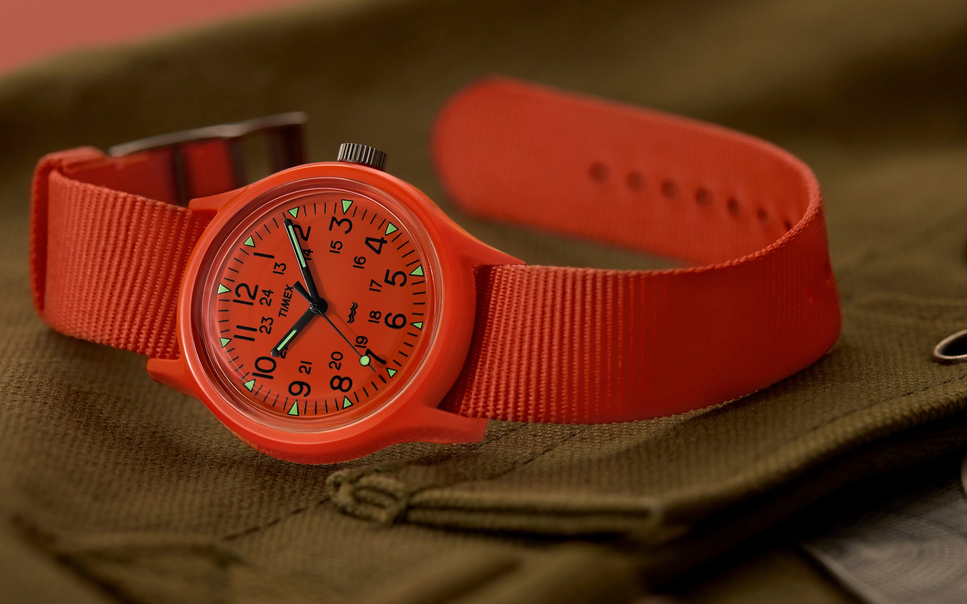 A Timex MK1 wristwatch with a bold red case and matching fabric strap is displayed on an olive-green fabric surface. The watch face features black Arabic numerals for both 12-hour and 24-hour time formats, with luminous triangular markers and hands. The black hour and minute hands contrast against the red dial, and a small wave symbol is present above the 6 o’clock position.