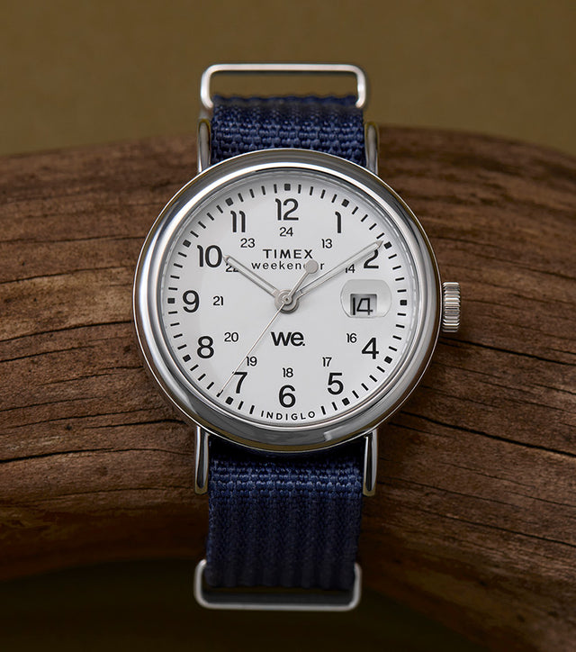 The Timex Weekender 37mm watch resting on a curved driftwood piece, featuring a white dial, black Arabic numerals, and a navy-blue fabric strap.