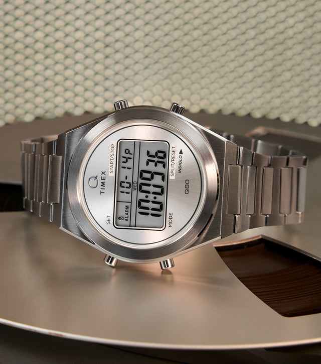 A gold-tone Q Timex® Q80 Continental digital watch with a round case and metal bracelet, displayed on a coiled blue telephone cord atop a white fabric surface. The digital display shows the time, day, and date.