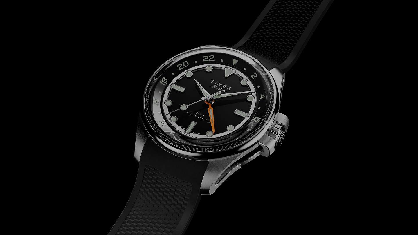 The Timex Atelier GMT Automatic watch with a black dial, orange GMT hand, and black rubber strap displayed against a black background.