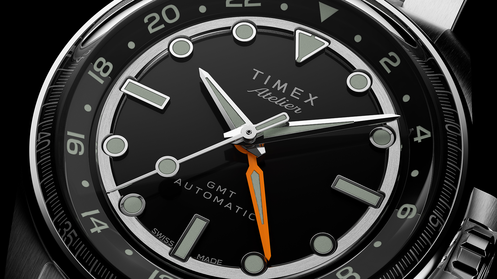 A close-up of the Timex Atelier GMT Automatic dial, highlighting the luminous markers, orange GMT hand, and polished silver hands.