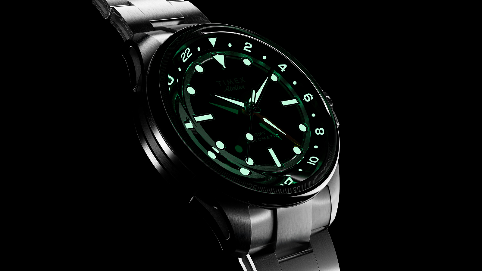 The Timex Atelier GMT Automatic watch glowing in the dark, showing luminous hour markers and hands in green light.