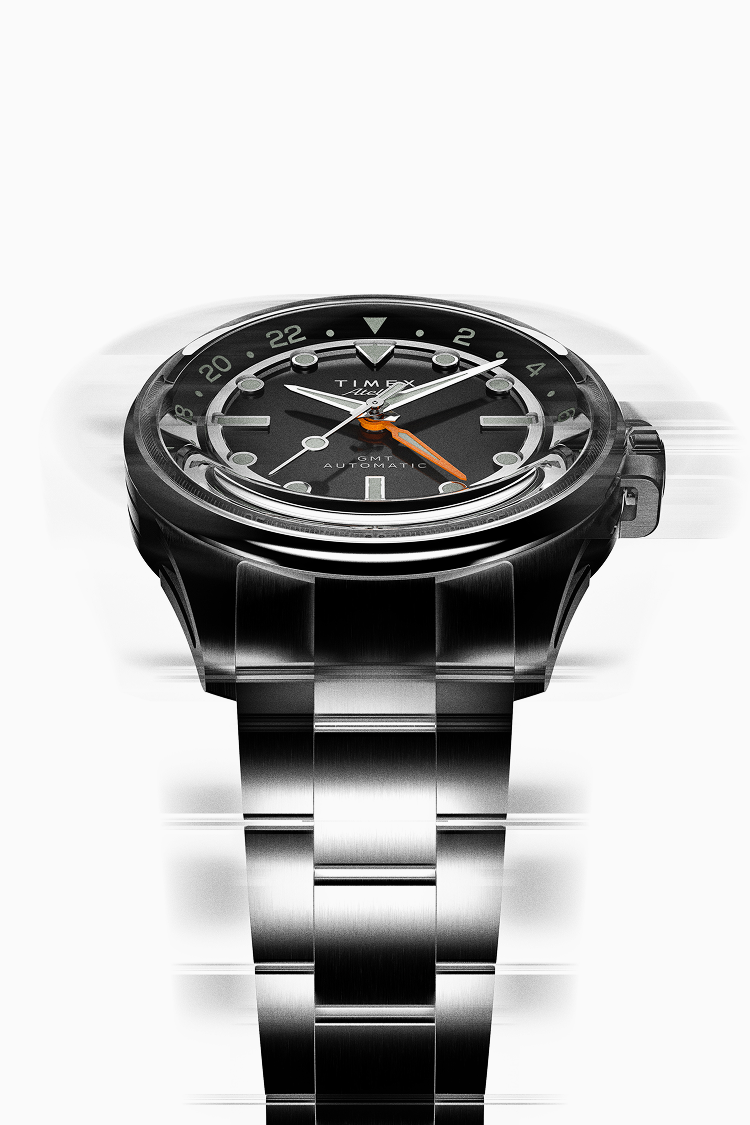 Front view of the Timex Atelier GMT Automatic highlighting its black dial, orange GMT hand, and brushed steel bracelet with a motion blur effect.