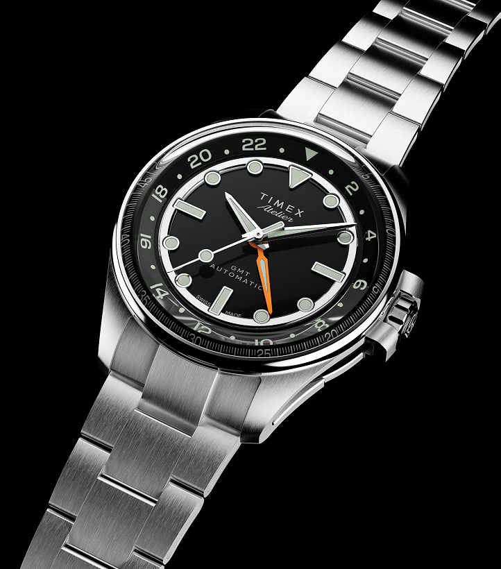 The Timex Atelier GMT Automatic with a stainless steel bracelet and black dial, featuring a bold orange GMT hand, shot from an angled perspective.