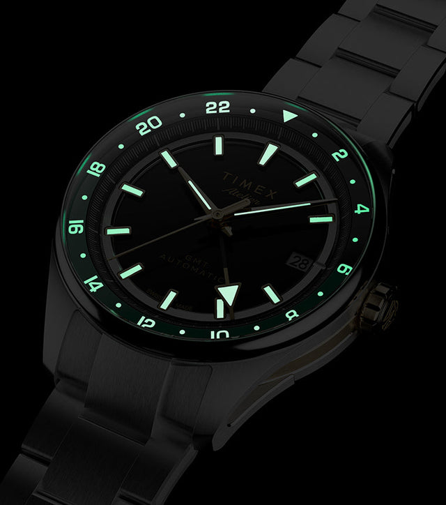 Timex Atelier GMT Première M1a watch glowing in the dark with illuminated hour markers, hands, and 24-hour scale.