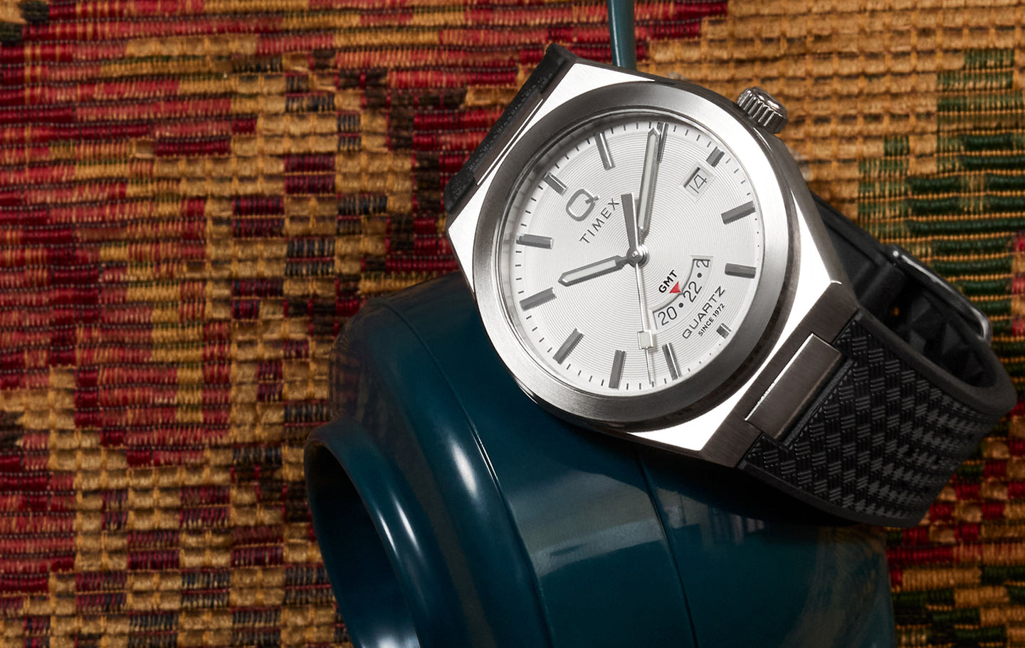 Q Timex® Continental GMT watch with white dial, stainless steel case, and black textured strap displayed on a blue ceramic vase against a woven textile background.