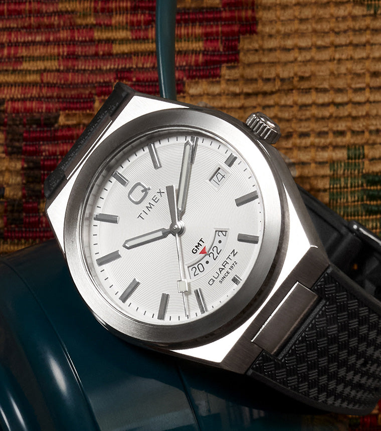 Q Timex® Continental GMT watch with white dial, stainless steel case, and black textured strap displayed on a blue ceramic vase against a woven textile background.