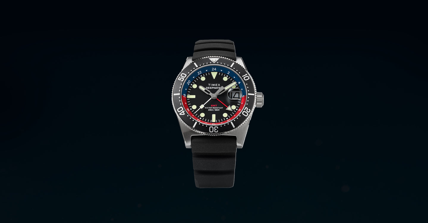 Timex Deepwater Reef 200 GMT dive watch is set against a dark underwater background. The watch features a stainless steel case, a black dial with luminous markers, and a blue and red GMT bezel. The bold hands and date window at 3 o’clock enhance functionality, while the black rubber strap adds to its rugged, water-resistant design. The watch is labeled “200M Water Resistant”, emphasizing its durability for underwater exploration.