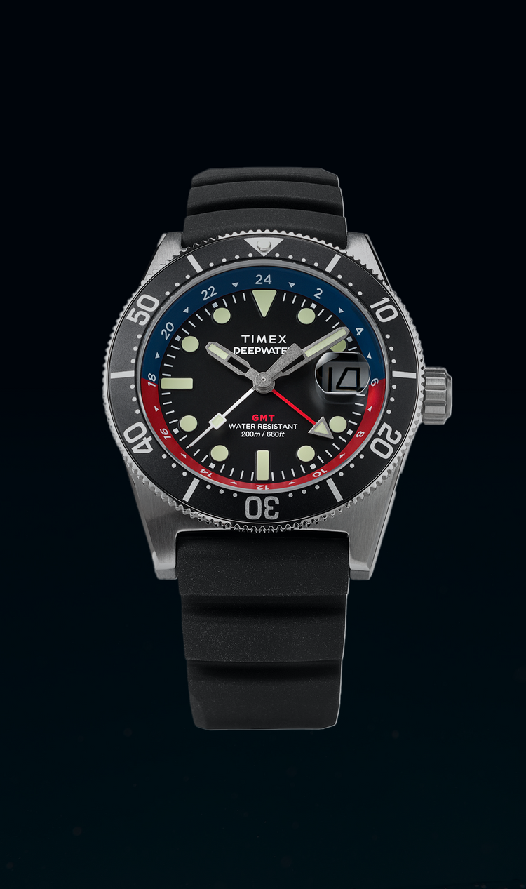 Timex Deepwater Reef 200 GMT dive watch is set against a dark underwater background. The watch features a stainless steel case, a black dial with luminous markers, and a blue and red GMT bezel. The bold hands and date window at 3 o’clock enhance functionality, while the black rubber strap adds to its rugged, water-resistant design. The watch is labeled “200M Water Resistant”, emphasizing its durability for underwater exploration.