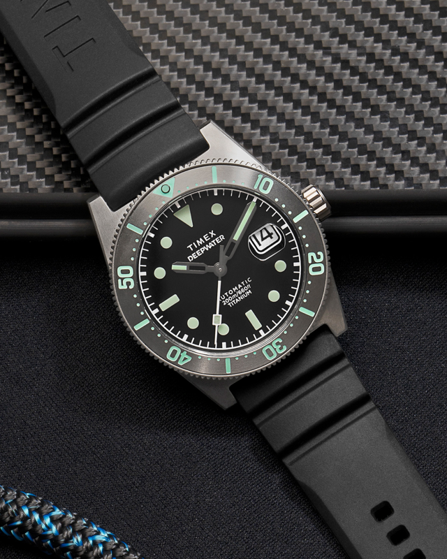 Image of the Timex Deepwater Reef 200 Titanium Automatic dive watch. The watch features a matte black dial with luminous markers, a unidirectional rotating bezel with green accents, and a date window at 3 o’clock with a magnifier. The titanium case and black rubber strap emphasize durability and lightweight comfort. The watch rests on a textured carbon fiber surface, with a coiled black and blue rope nearby, reinforcing its rugged, adventure-ready design.