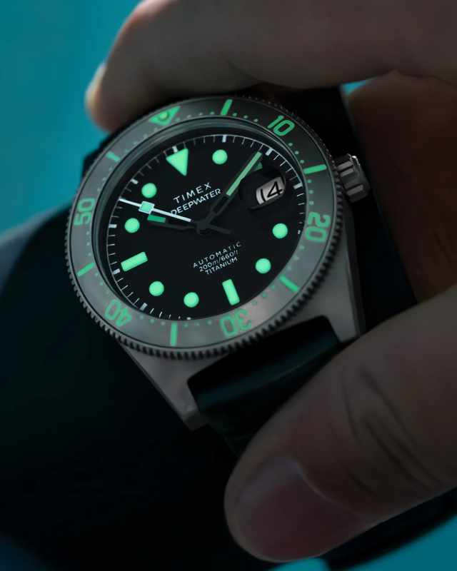 Image of the Timex Deepwater Reef 200 Titanium Automatic dive watch in low-light conditions, showcasing its glowing luminous markers and bezel accents. The watch features a black dial, a unidirectional rotating bezel, a date window with a magnifier, and a durable titanium case. A hand grips the watch, emphasizing its rugged and adventure-ready design. The background has a soft blue aquatic tone, reinforcing its deep-sea functionality and water resistance up to 200 meters.