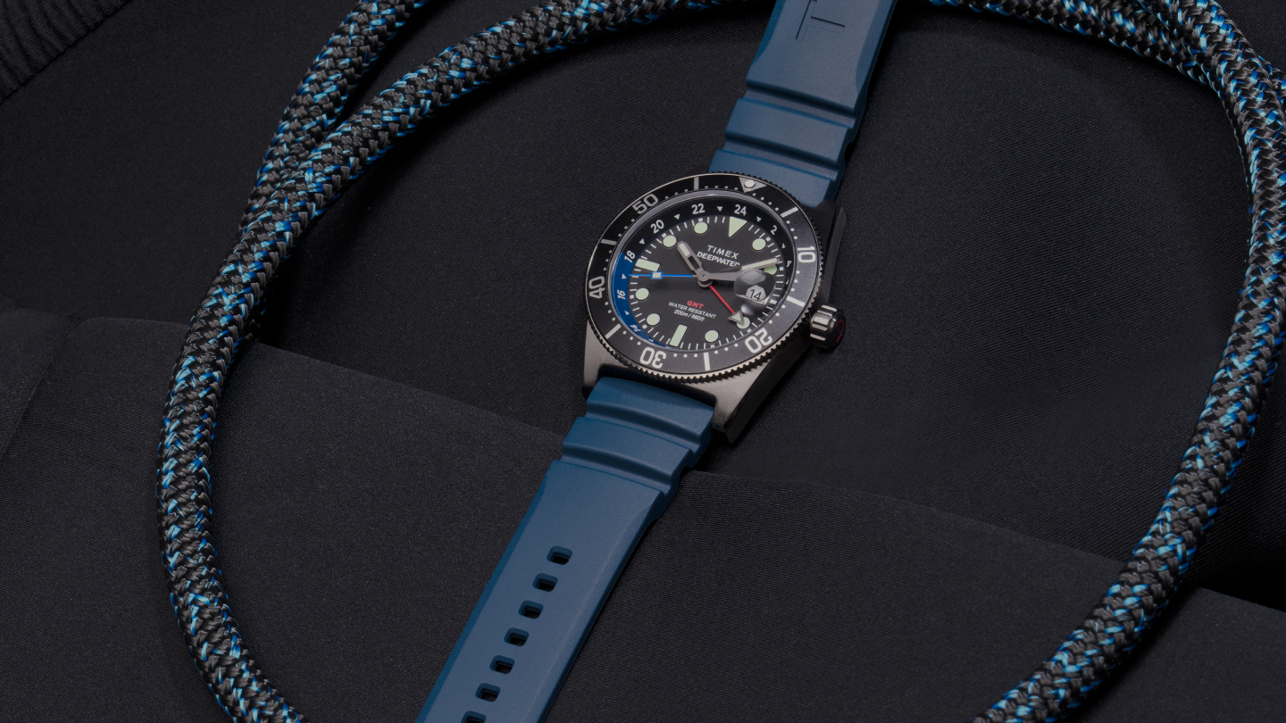 Image of the Timex Deepwater Reef 200 GMT dive watch with a blue silicone strap, placed on a black wetsuit. The watch features a black dial with luminous markers, a GMT hand, a rotating bezel with blue and black accents, and a date window at 3 o’clock. A coiled black and blue rope surrounds the watch, reinforcing its rugged, adventure-ready design. The composition highlights the watch’s durability and water-resistant functionality, making it ideal for diving and exploration.