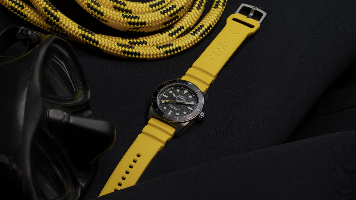 Image of the Timex Deepwater Reef 200 dive watch with a bold yellow silicone strap, placed on a black wetsuit. The watch features a textured black dial, luminous markers, a unidirectional rotating bezel, and a rugged case. A black diving mask and a coiled yellow and black rope surround the watch, emphasizing its durability and adventure-ready design. The composition highlights the watch’s water-resistant functionality, ideal for diving and exploration.