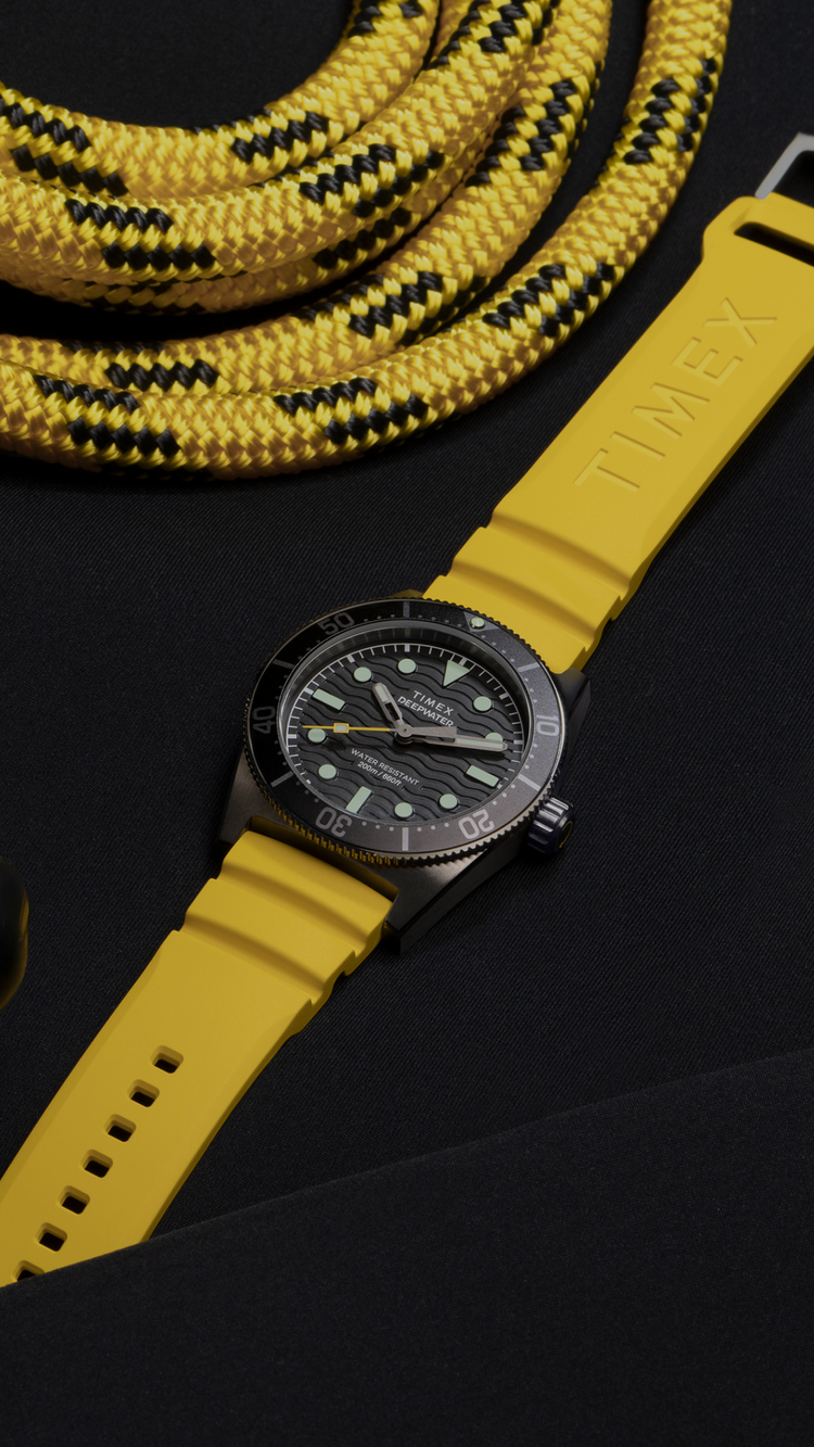 Image of the Timex Deepwater Reef 200 dive watch with a bold yellow silicone strap, placed on a black wetsuit. The watch features a textured black dial, luminous markers, a unidirectional rotating bezel, and a rugged case. A black diving mask and a coiled yellow and black rope surround the watch, emphasizing its durability and adventure-ready design. The composition highlights the watch’s water-resistant functionality, ideal for diving and exploration.