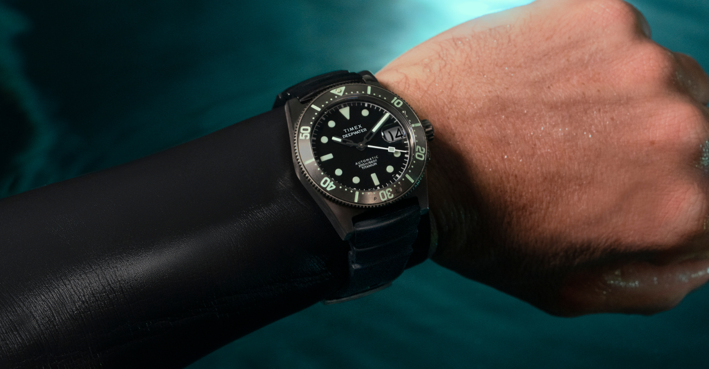 Timex Deepwater Reef 200 Titanium Automatic dive watch worn over a black wetsuit sleeve. The watch features a black dial, luminous markers, a rotating bezel with green accents, and a titanium case. The background shows a blurred aquatic environment, highlighting the watch’s rugged, water-resistant design for underwater exploration.