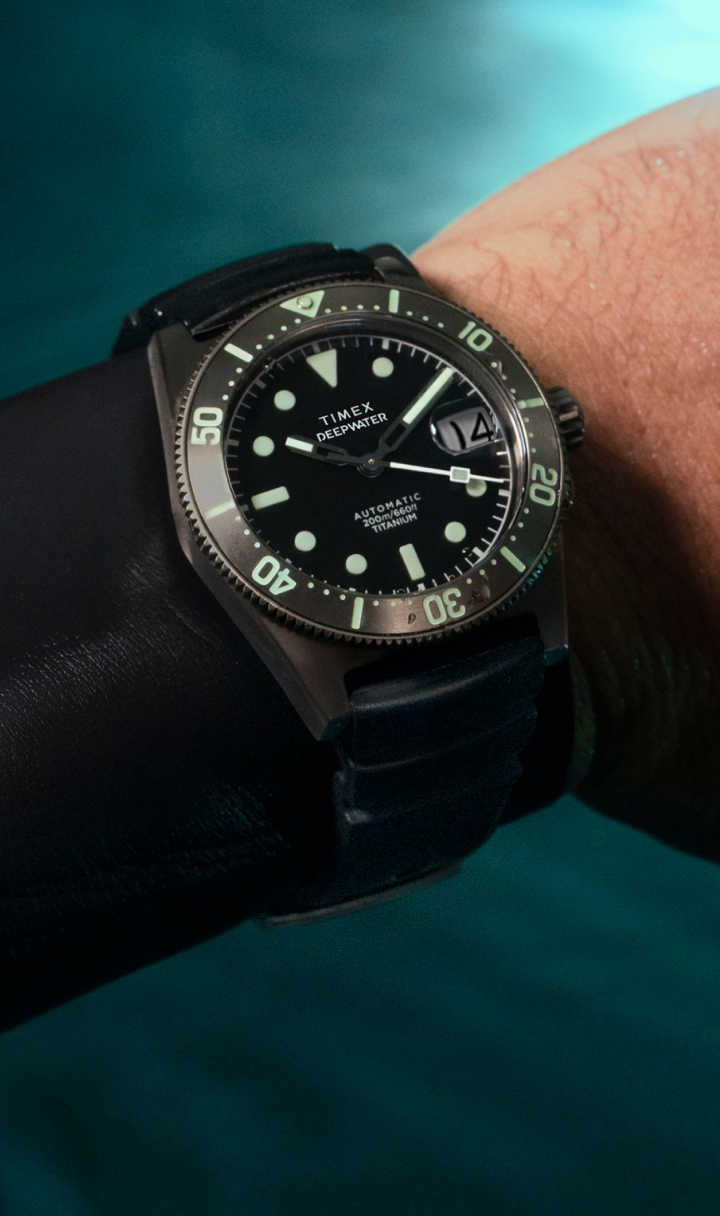 Timex Deepwater Reef 200 Titanium Automatic dive watch worn over a black wetsuit sleeve. The watch features a black dial, luminous markers, a rotating bezel with green accents, and a titanium case. The background shows a blurred aquatic environment, highlighting the watch’s rugged, water-resistant design for underwater exploration.