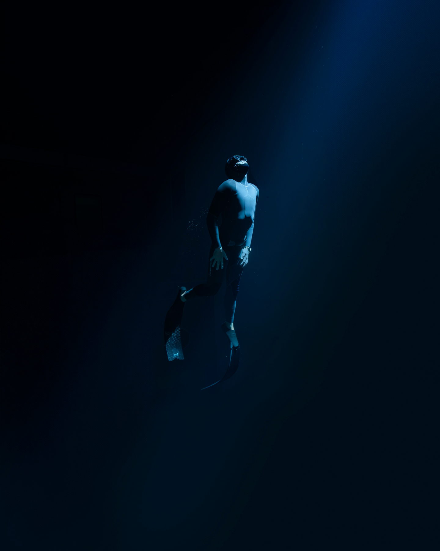 A deep underwater scene shows a diver in a black wetsuit and mask, floating in a dimly lit environment. A soft blue light from above casts a glow on the diver, illuminating small air bubbles escaping from their mask. The diver's posture is relaxed, head tilted back as if gazing upward. The surrounding water is dark, creating a sense of depth and isolation.