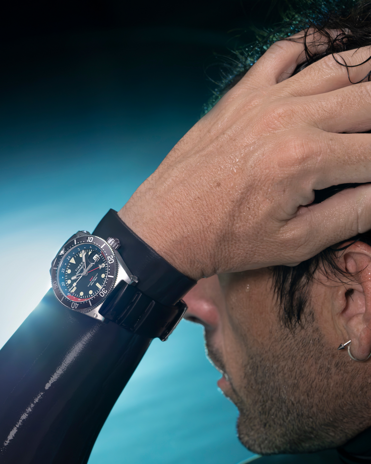 A close-up image of a man wearing the Timex Deepwater Reef 200 GMT dive watch. His wet hand is placed on his head, with damp hair and beads of water visible on his skin, suggesting recent underwater activity. The watch, strapped over a black wetsuit, features a black dial, luminous markers, a rotating bezel, and a durable rubber strap. The background is softly blurred with a blue aquatic tone, emphasizing the watch’s rugged, water-resistant design for diving and adventure.