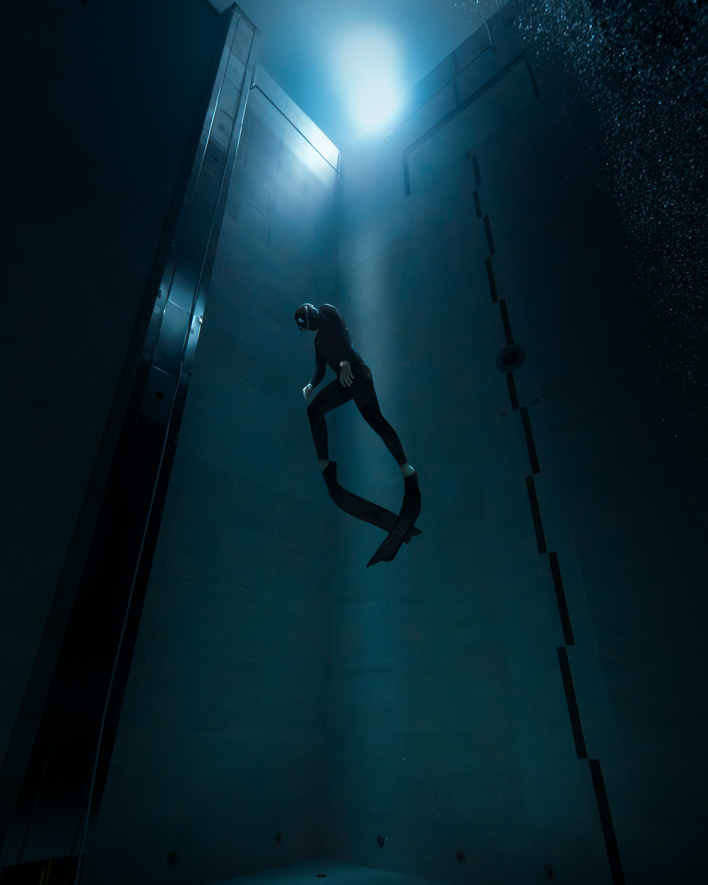 A deep underwater scene shows a diver in a black wetsuit and mask, floating in a dimly lit environment. A soft blue light from above casts a glow on the diver, illuminating small air bubbles escaping from their mask. The diver's posture is relaxed, head tilted back as if gazing upward. The surrounding water is dark, creating a sense of depth and isolation.