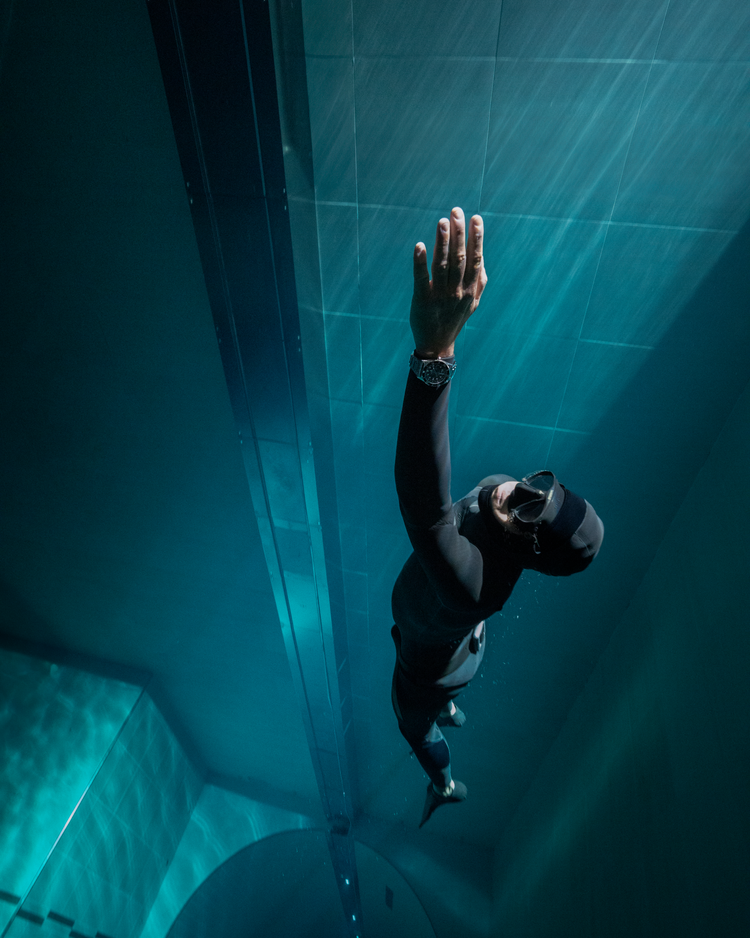 A dark underwater scene captures the lower half of a diver wearing a wetsuit and long black fins. The diver's feet are partially visible, with one foot slightly exposed from the fin. The surrounding water is deep and shadowy, with minimal lighting creating an eerie, isolated atmosphere