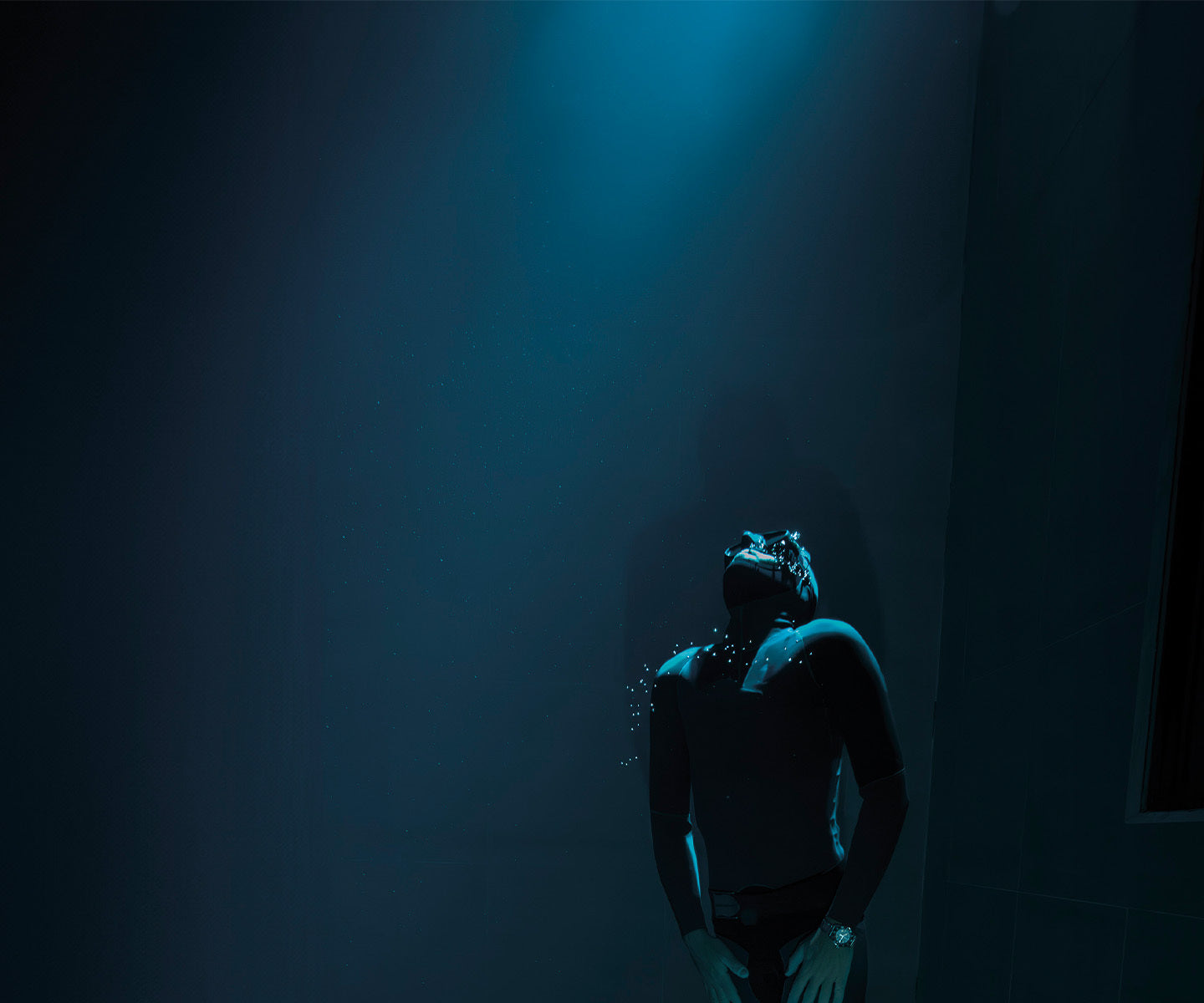 A deep underwater scene shows a diver in a black wetsuit and mask, floating in a dimly lit environment. A soft blue light from above casts a glow on the diver, illuminating small air bubbles escaping from their mask. The diver's posture is relaxed, head tilted back as if gazing upward. The surrounding water is dark, creating a sense of depth and isolation.