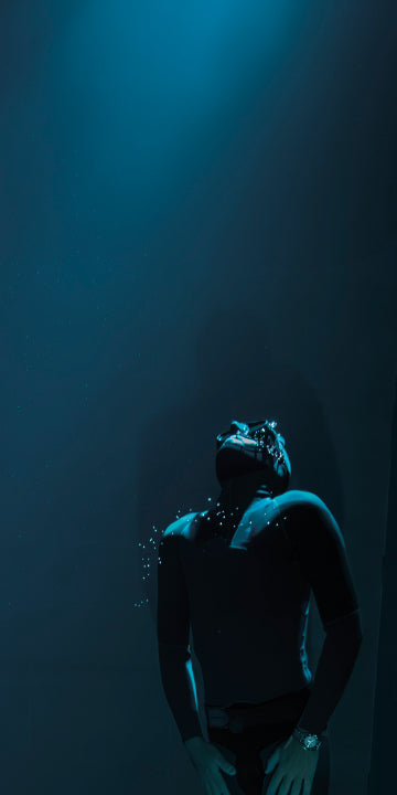 A deep underwater scene shows a diver in a black wetsuit and mask, floating in a dimly lit environment. A soft blue light from above casts a glow on the diver, illuminating small air bubbles escaping from their mask. The diver's posture is relaxed, head tilted back as if gazing upward. The surrounding water is dark, creating a sense of depth and isolation.