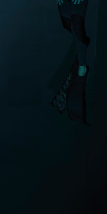 A dark underwater scene captures the lower half of a diver wearing a wetsuit and long black fins. The diver's feet are partially visible, with one foot slightly exposed from the fin. The surrounding water is deep and shadowy, with minimal lighting creating an eerie, isolated atmosphere