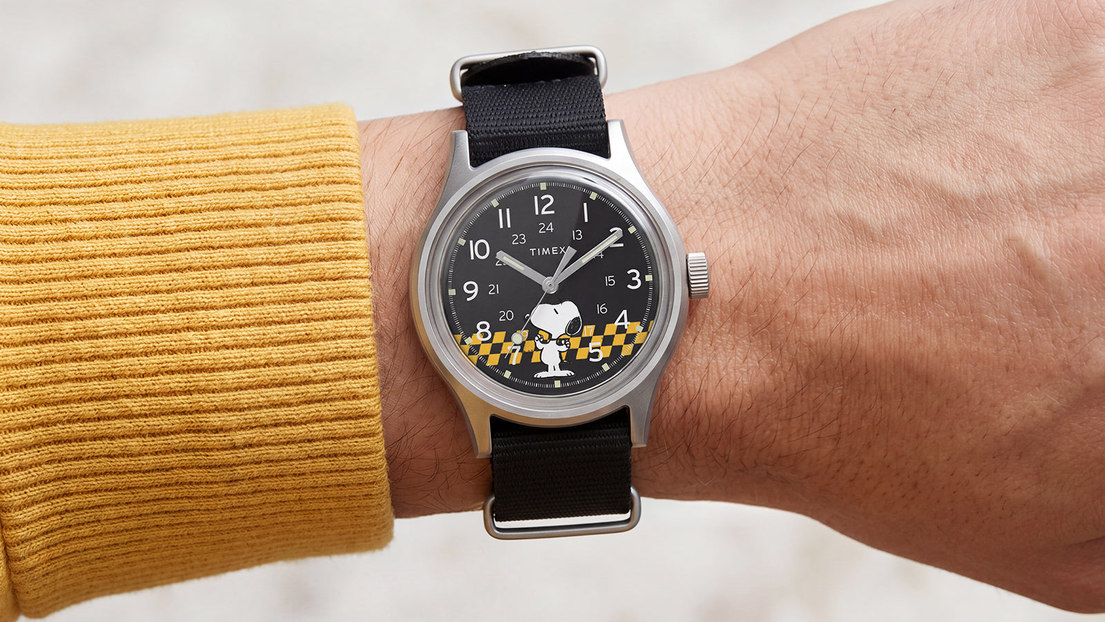 Peanuts® x Timex MK1® watch worn on the wrist, showcasing a black dial with Snoopy artwork, silver-tone case, and black fabric strap.