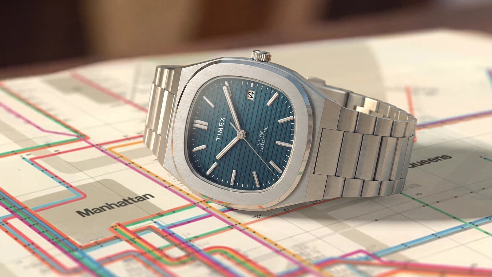 Timex Automatic E Line 38mm with a blue dial and stainless-steel bracelet, displayed across a colorful transit map.