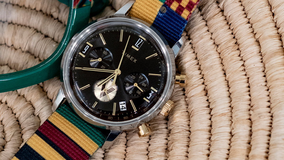 TWG069100 Close-up of the black-dial watch and colorful striped strap on a woven surface beside green glasses.