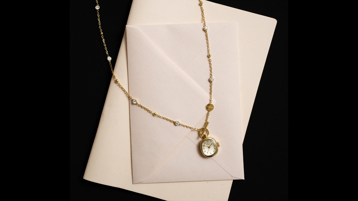 TW6A17700 Cavatina® Mini gold-tone watch necklace with clear stones, arranged across a cream envelope against a black background.