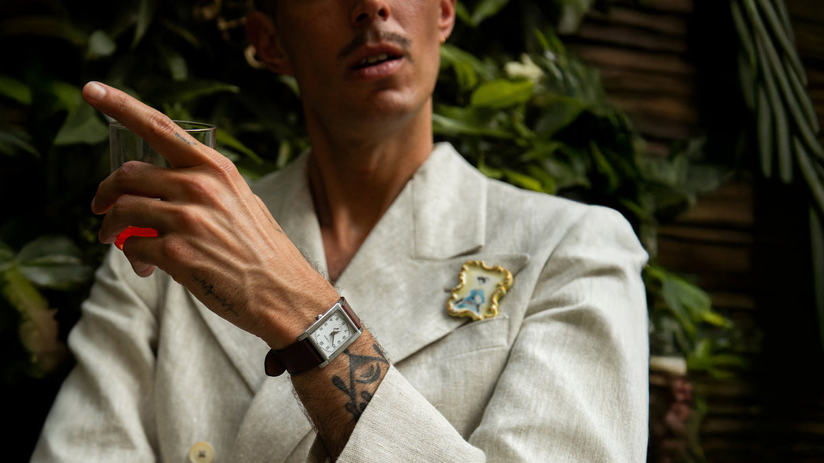 TWG069000 Model in a cream suit wearing a WM Brown x Timex watch with a brown leather strap while holding a drink.