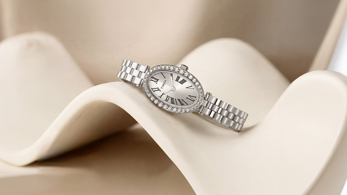 TW2Y90000 Silver-tone Timex Cavatina® Crystals watch arranged on a wave-shaped ivory display, featuring an oval dial and crystal-lined bezel.