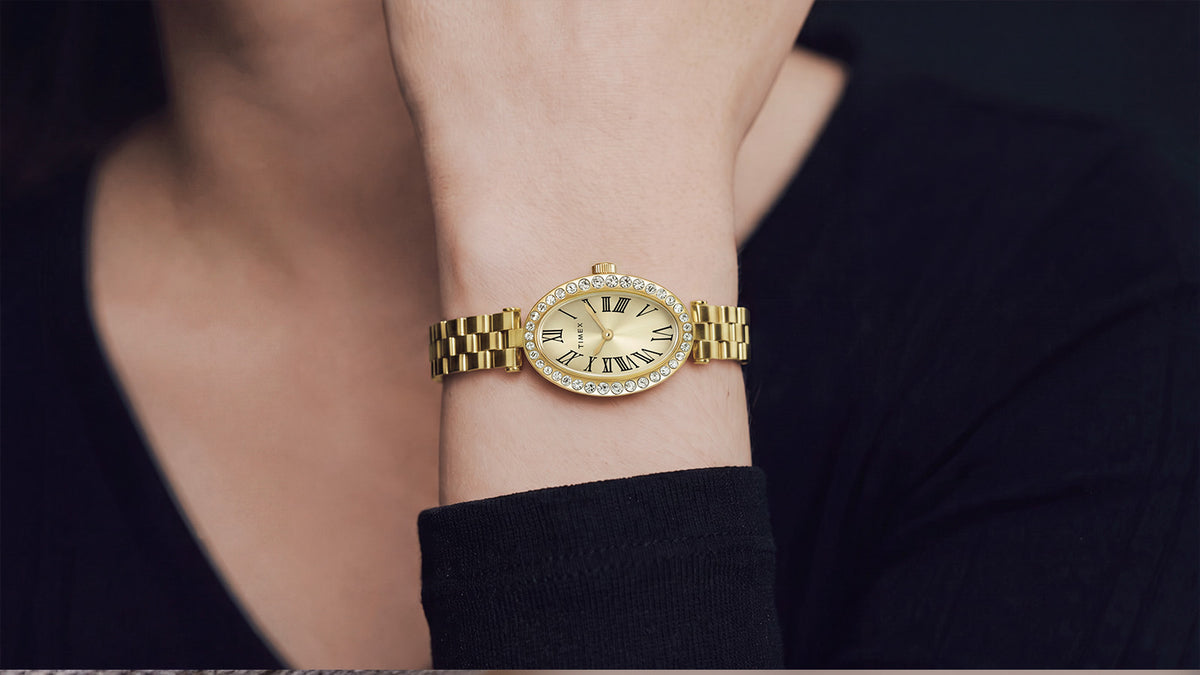TW2Y90100 Timex Cavatina® Crystals watch worn on a wrist, featuring a gold-tone link bracelet and crystal-lined oval case against a black outfit.