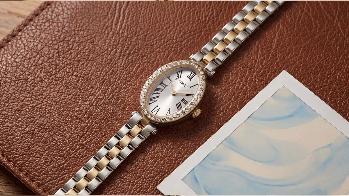 TW2Y90200 Timex Cavatina® Crystals watch with a silver-and-gold-tone link bracelet, resting diagonally on brown leather beside a photograph.