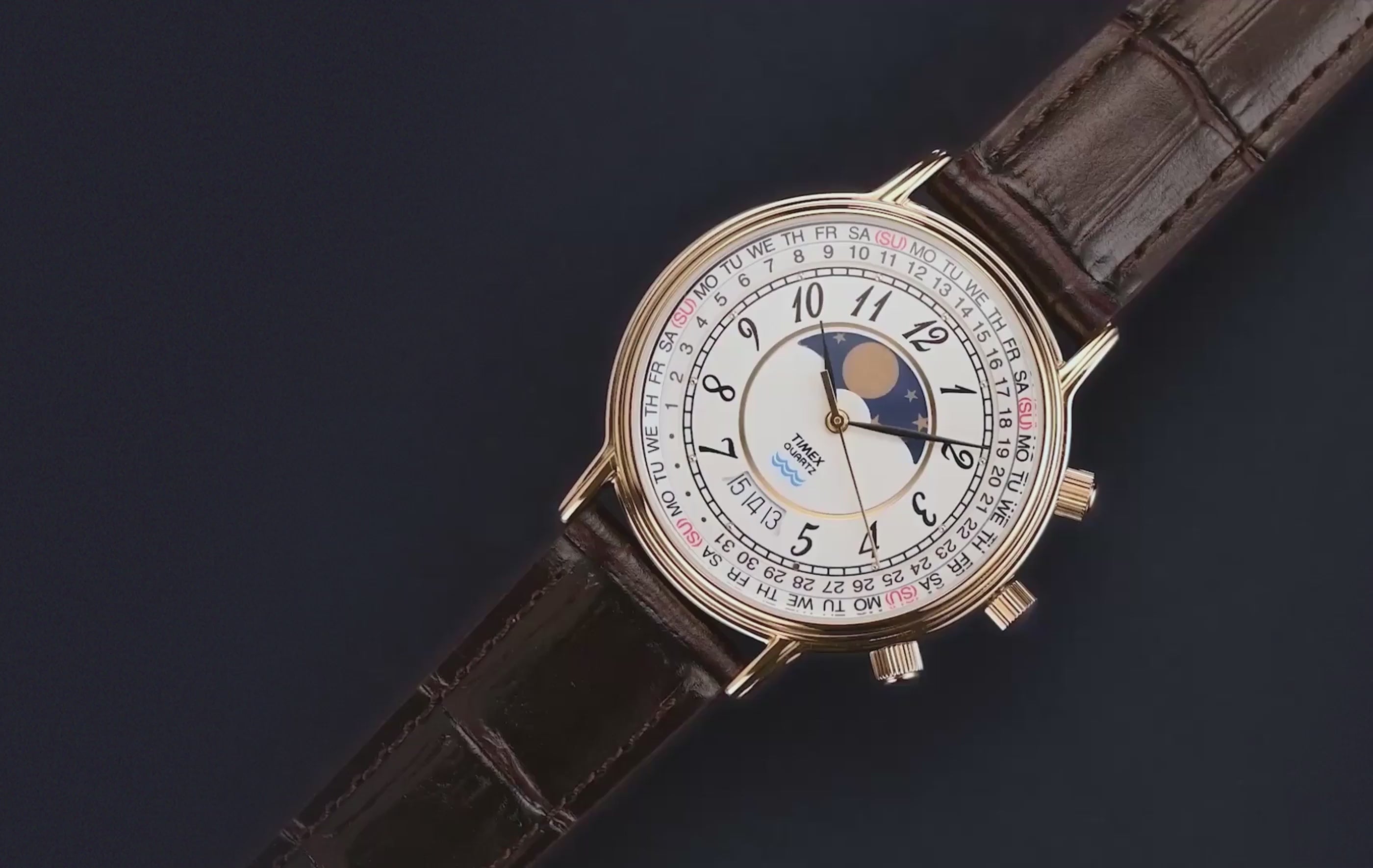 Timex 1990 Moon Phase and Calendar Dial Reissue watch with a gold-tone case, detailed white dial, and brown leather strap rotating against a dark background.