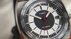 Video showcasing the Timex World Time Reissue watch (model TW2V69500) rotating on an orange leather surface. The footage highlights the brushed stainless-steel case, domed crystal, black-and-white dial with red seconds hand, and engraved world time bezel displaying city names such as New York, Tokyo, and Sydney.