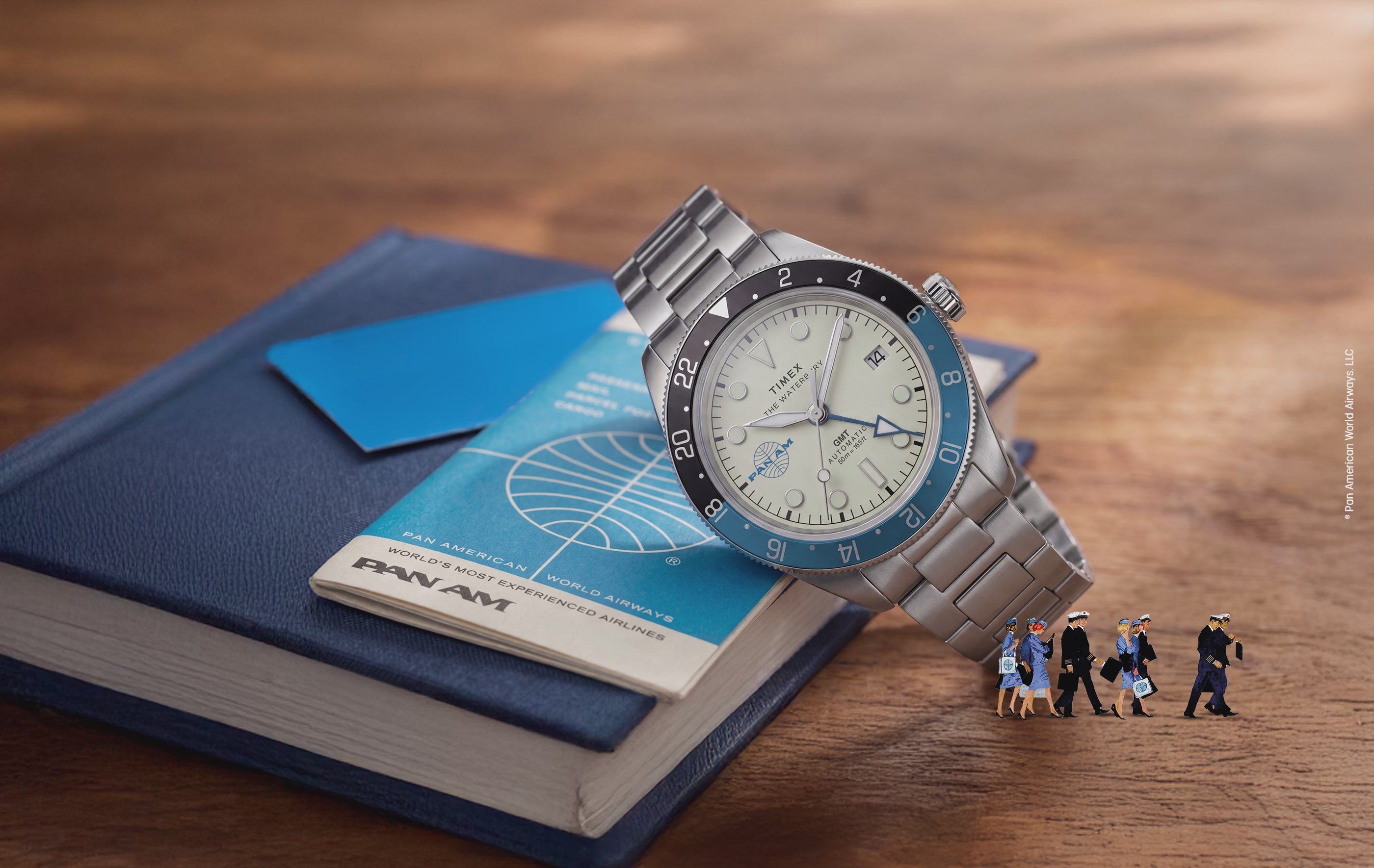 TW2Y94300 Pan Am® x Timex® Waterbury GMT watch with a cream dial, blue-and-black bezel and silver-tone bracelet resting on a Pan Am booklet, with miniature airline crew figures below and an animated airplane flying overhead.