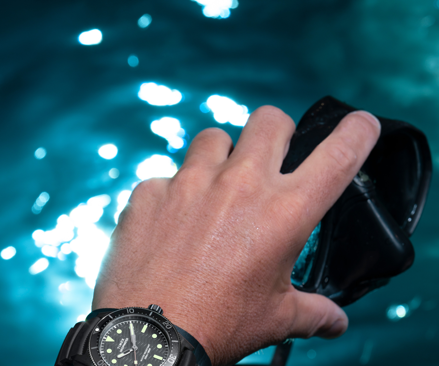 Image of a diver’s hand holding a black diving mask, with the Timex Deepwater Reef 200 dive watch on their wrist. The watch features a black dial, luminous markers, a rugged bezel, and a durable strap, emphasizing its water-resistant and adventure-ready design. The background showcases shimmering blue water with sunlight reflections, reinforcing the oceanic and diving theme.