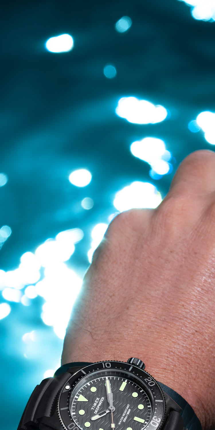 Image of a diver’s hand holding a black diving mask, with the Timex Deepwater Reef 200 dive watch on their wrist. The watch features a black dial, luminous markers, a rugged bezel, and a durable strap, emphasizing its water-resistant and adventure-ready design. The background showcases shimmering blue water with sunlight reflections, reinforcing the oceanic and diving theme.