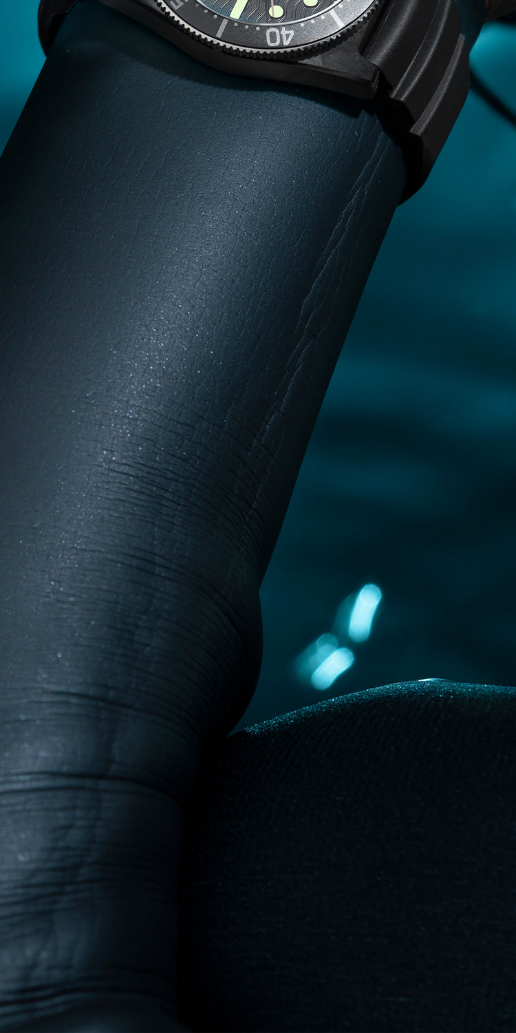 Image of a diver’s arm in a black wetsuit, partially submerged in water, with the Timex Deepwater Reef 200 dive watch visible on the wrist. The background features deep blue water with shimmering light reflections, creating a moody and immersive underwater atmosphere. The composition emphasizes the watch’s durability and functionality for aquatic adventures.