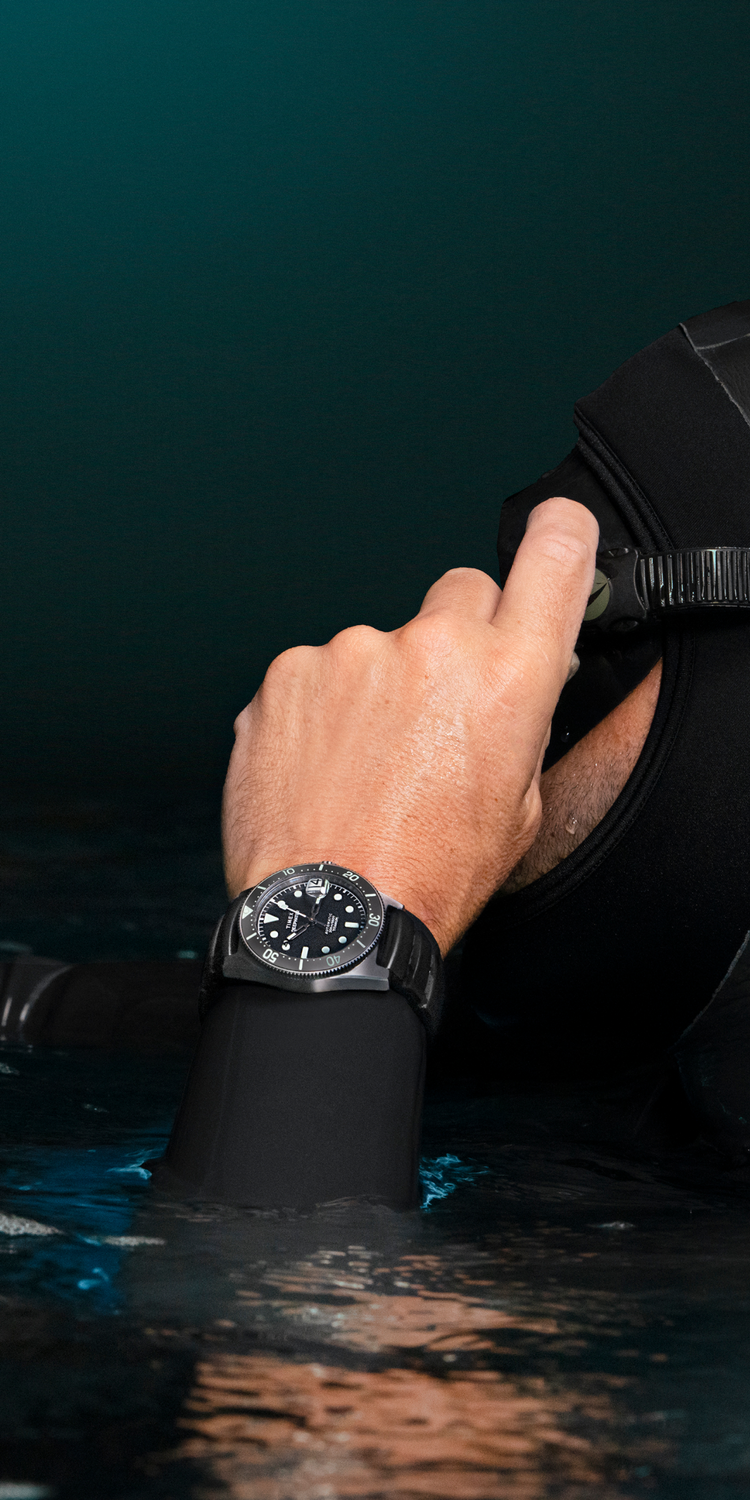 A diver wearing a black wetsuit and diving hood adjusts their mask while partially submerged in dark waters. On their wrist is the Timex Deepwater Reef 200 Titanium Automatic dive watch, featuring a black dial, luminous markers, and a rugged titanium case. The scene is illuminated by subtle ambient light, highlighting the watch’s durability and water resistance. The composition emphasizes the watch as a reliable tool for underwater exploration.